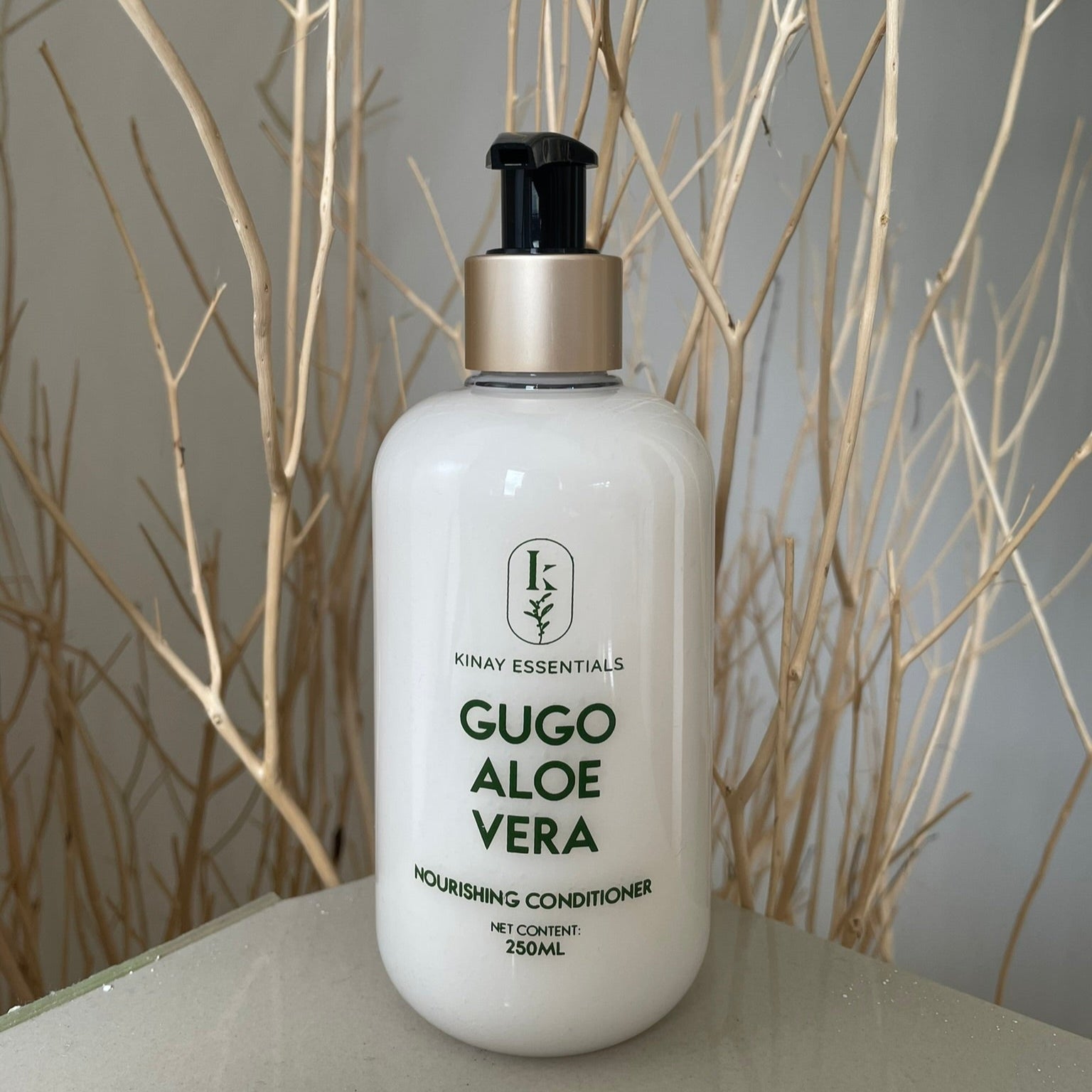 White bottle of Gugo Aloe Vera conditioner with a black cap on a gray surface with bare branches in the background.