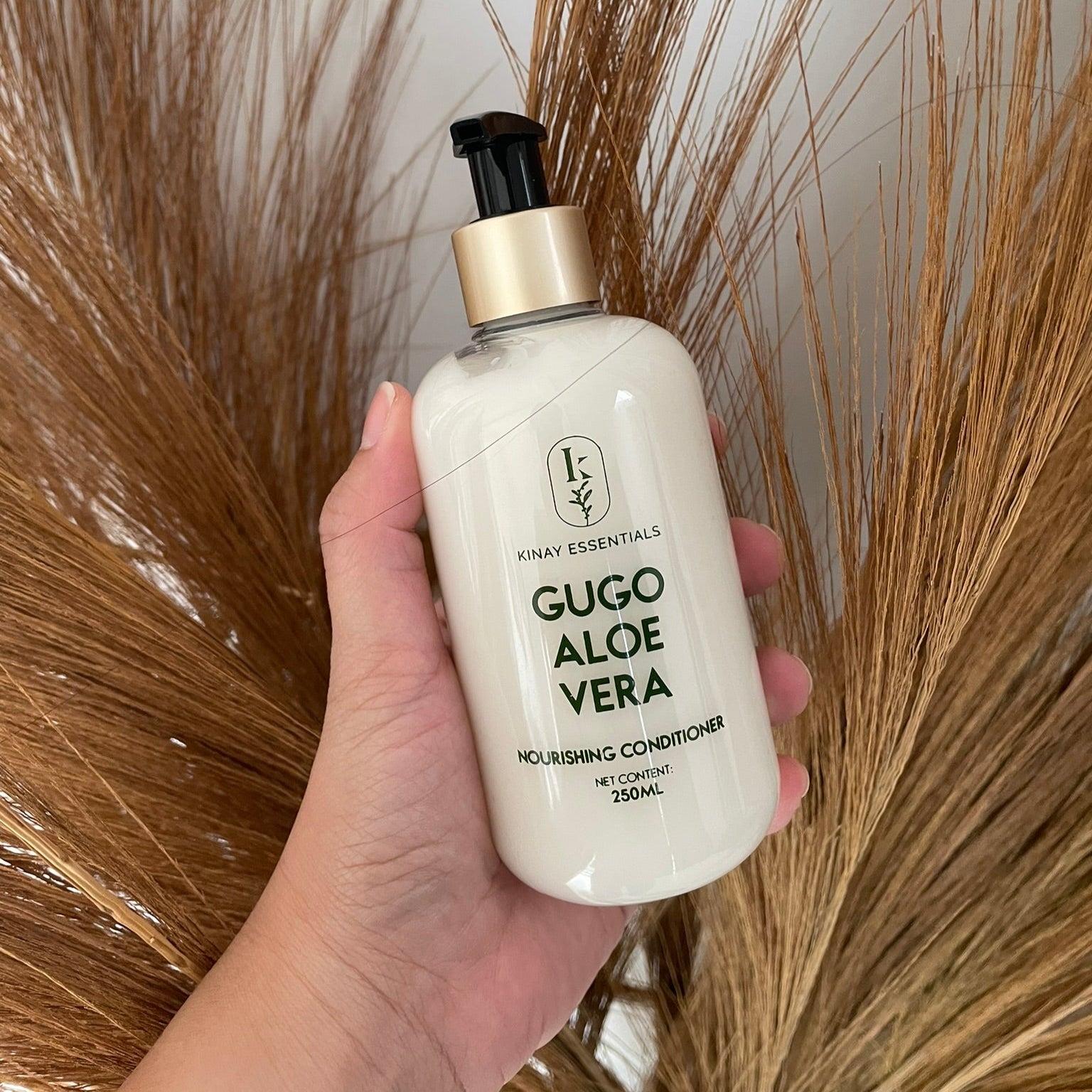 Hand holding a bottle of 'Gugo Aloe Vera' conditioner against a background of dried pampas grass.