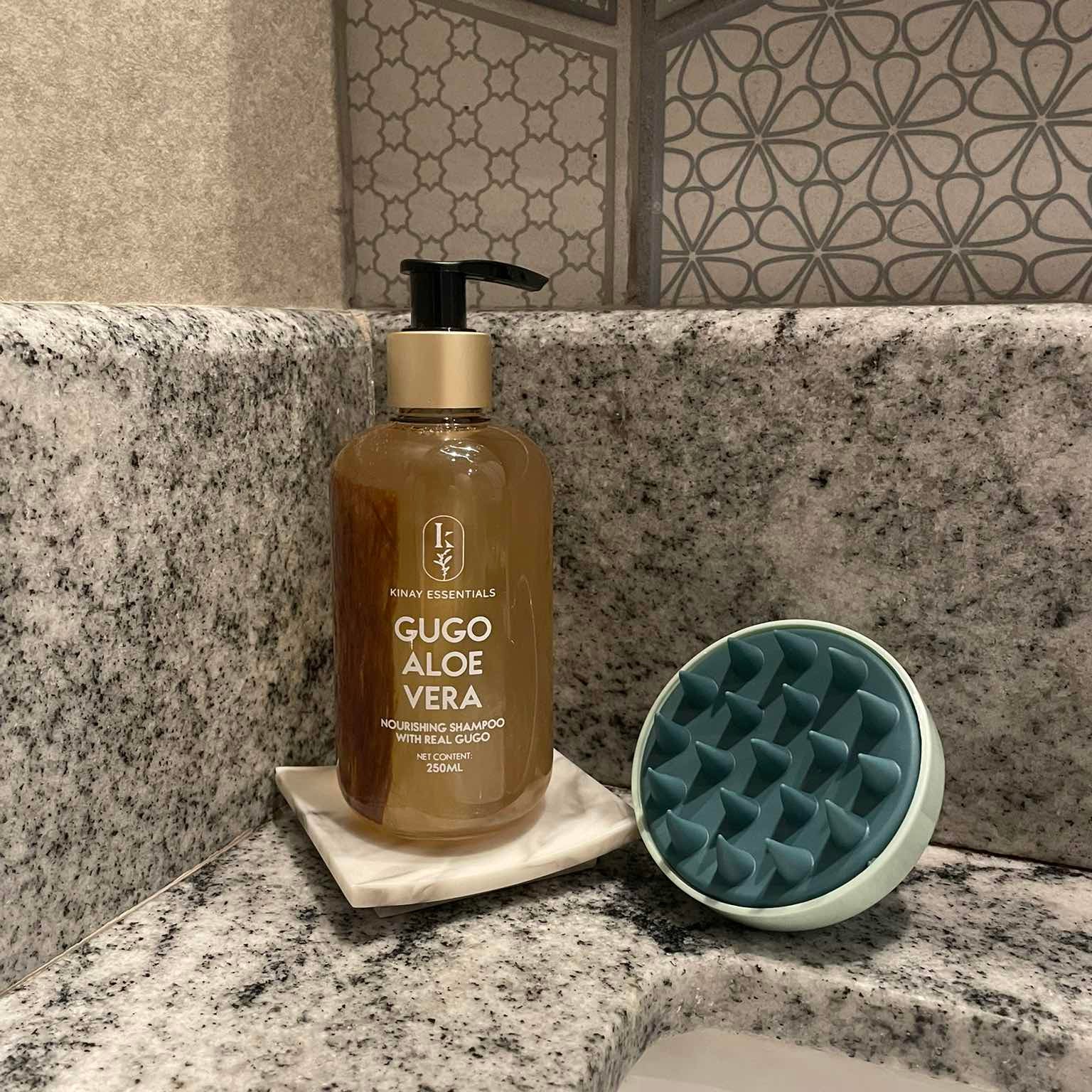 Bottle of kinay essentials Gugo Aloe Vera shampoo on a bathroom counter with a scalp massager beside it