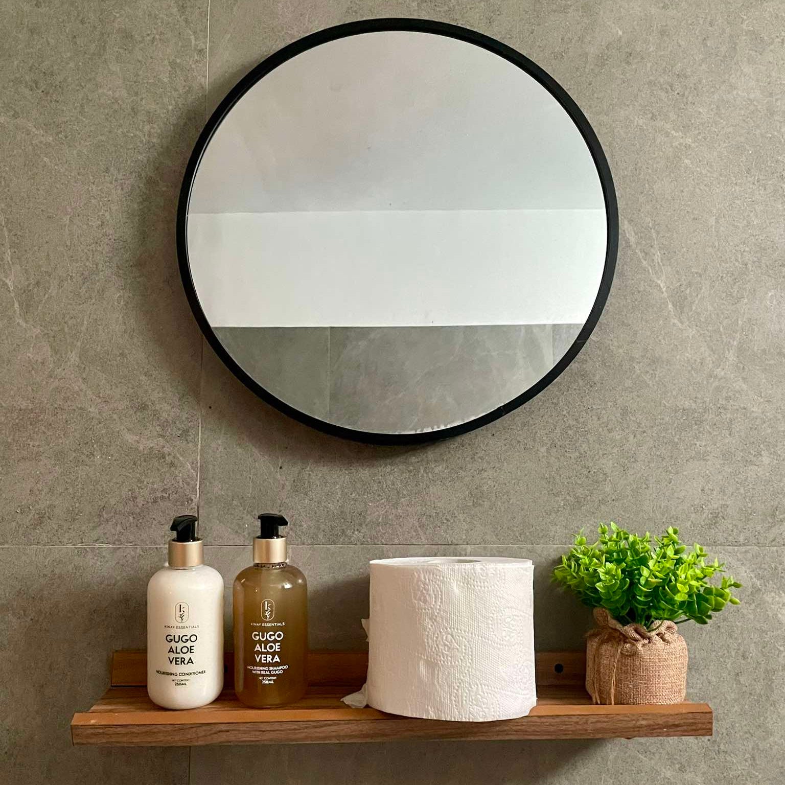 Round mirror with a wooden shelf holding bottles of gugo aloe vera shampoo & conditioner and a plant on a gray tiled wall.