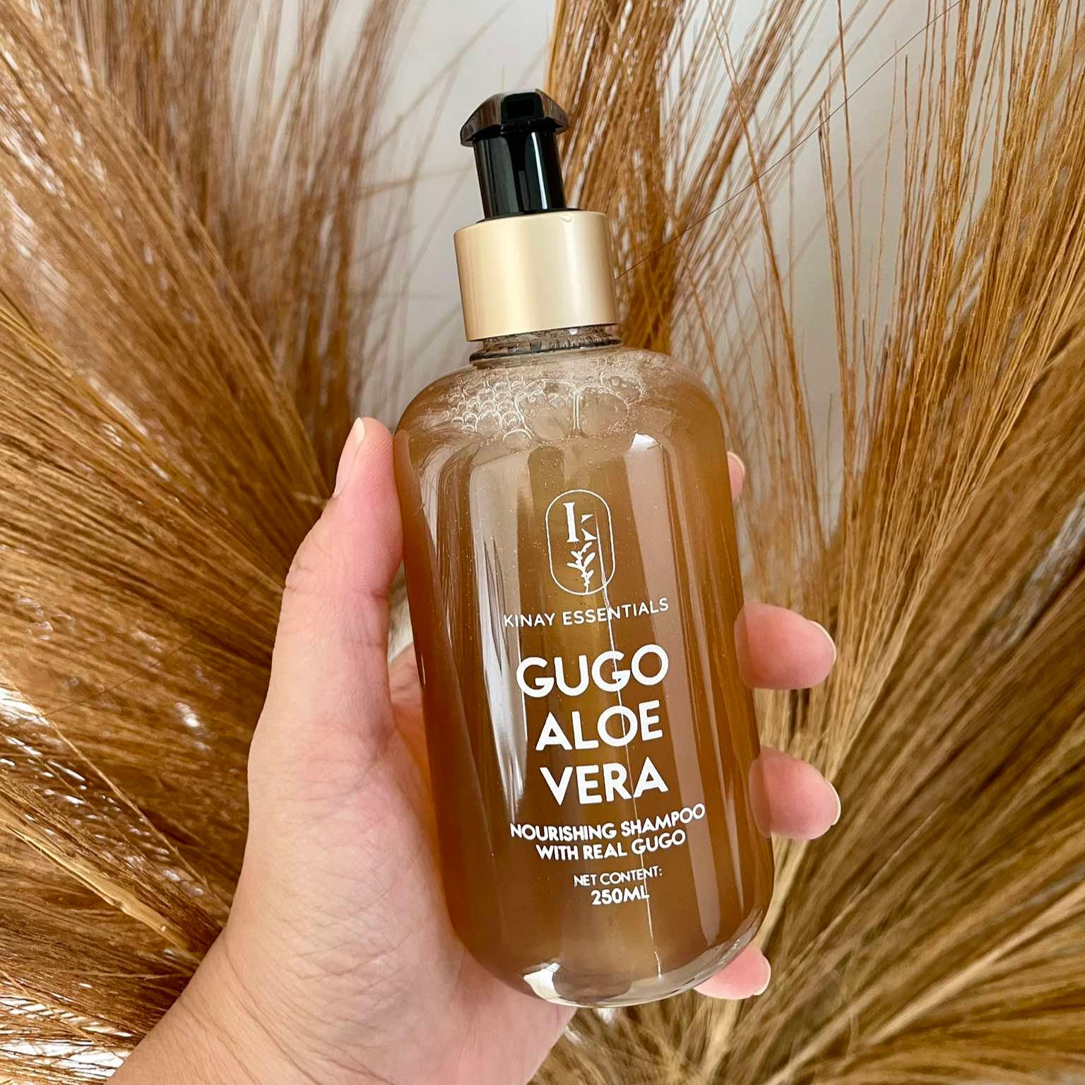 Hand holding a bottle of kinay essentials   Gugo Aloe Vera shampoo with a dried plant background