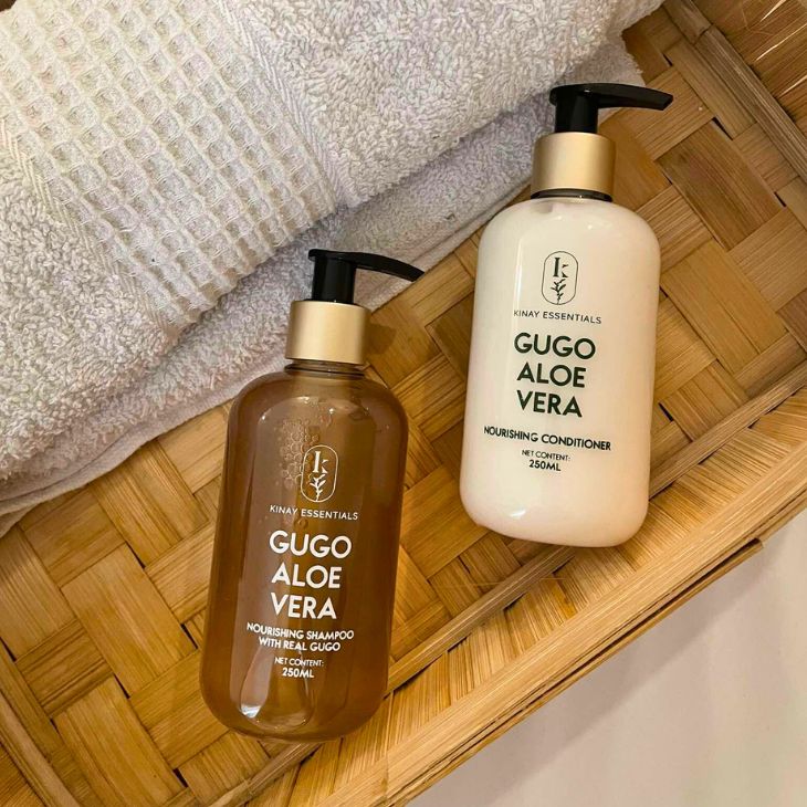 Two bottles of Gugo Aloe Vera shampoo and conditioner on a woven mat with a towel in the background.