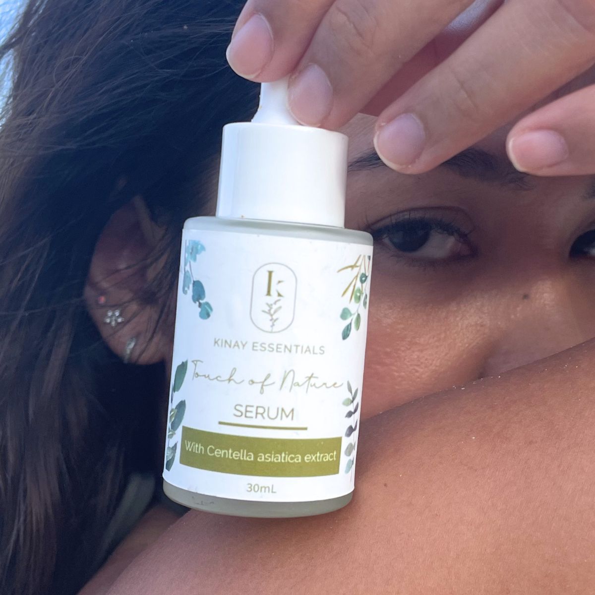 Person holding a bottle of 'Touch of Nature Serum' by Kinay Essentials close to their face.